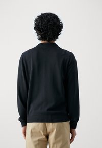Person mit lockigem schwarzem Haar, der einen schwarzen Pullover und beiges Hosen trägt, von hinten vor einem schlichten weißen Hintergrund gezeigt.