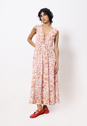 Robe maxi blanche avec des motifs floraux et d'objets rouges complexes, un décolleté en V profond, des manches courtes, et une taille froncée, associée à des ballerines rouges.