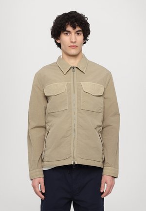 Giacca beige chiaro con zip frontale, due tasche sul petto e tasche laterali. Ha una vestibilità rilassata e un tessuto liscio, leggermente strutturato.