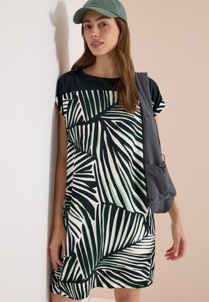 Femme portant une casquette verte et une robe noire avec des motifs de feuilles blanches et vertes, portant un sac à épaule gris, adossée à un mur.