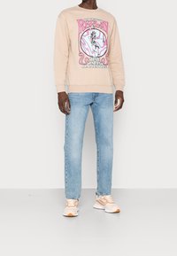 Felpa beige con una stampa grafica colorata, abbinata a jeans azzurri chiari e sneakers beige con dettagli arancioni.