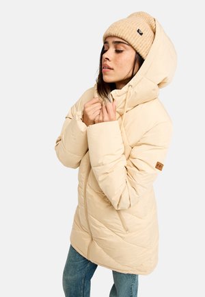 Femme portant une doudoune beige avec capuche et un bonnet en maille assorti, debout les yeux fermés et les mains près du col, sur un fond uni.