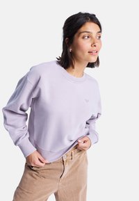 Felpa lavanda in tessuto morbido con colletto a girocollo e polsini a costina. Presenta un piccolo logo sul petto, abbinata a pantaloni in velluto a coste color beige.