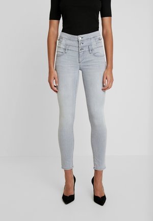 Kvinde iført lysegrå højtaljede skinny jeans med treknapslukning og sorte spidse hæle.
