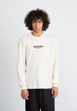 Light beige ribbed long-sleeve shirt with a black "Dickies Since '22" logo on the front. Crew neck and relaxed fit. Paired with dark pants.