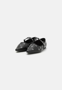 Ballerines en cuir noir avec des bouts pointus, présentant des accents en œillets blancs et une sangle croisée au-dessus du coup de pied. Texture lisse, design minimaliste.