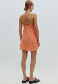 Orangfarbenes Kleid mit einem lockeren Schnitt und dünnen Trägern, das ein tief ausgeschnittenes Rückendesign hat und aus weichem Stoff gefertigt ist. Das Model trägt schwarze Plateausandalen.