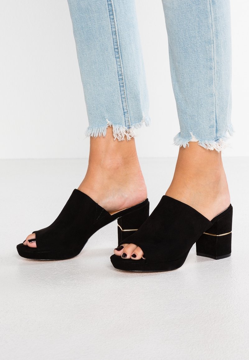 Pieds portant des mules à talons blocs en suède noir à bout ouvert, associées à un jean bleu clair avec ourlet effiloché, sur une surface blanche.