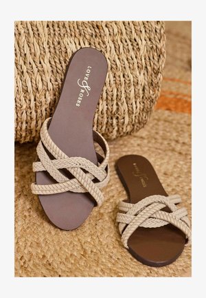 Geweven beige sandalen met een platte zool, met een donkerbruine binnenzool en een logo in goud. Het gestructureerde bandontwerp geeft een geweven uitstraling.