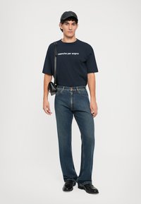 Marinblå bomullströja med vit text, kombinerad med blå raka jeans. Tillbehör inkluderar en svart keps och en liten svart axelväska.