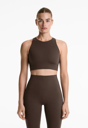 A nő felsőtesten előrenéz, sötétbarna ujjatlan cropped felsőt és magasan derekú leggingset visel egy sima fehér háttér előtt.