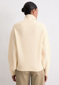 Crèmewitte, geribbelde gebreide trui met een hoge kraag en verzakte schouders, gecombineerd met beige pantalon. Zachte, gestructureerde stof.