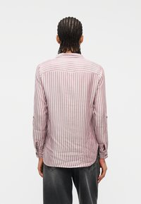 Άτομο με πλεγμένα μαλλιά, που φοράει ένα ροζ και λευκό πουκάμισο με κάθετες ρίγες μακρυμάνικο και σκουροπλυμένα τζιν, γυρισμένο πλάτη στην κάμερα.