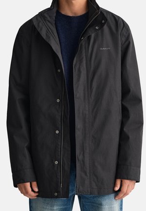 Mężczyzna ubrany w czarną kurtkę na zamek nałożoną na ciemnoniebieski sweter i niebieskie jeansy, kurtka z logo "GANT" na piersi.