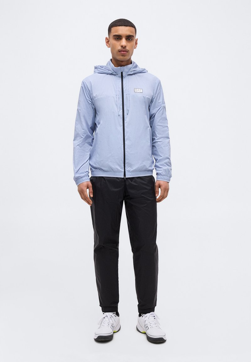 Νέος άνδρας που στέκεται με το πρόσωπο προς τα εμπρός, φοράει ανοιχτό μπλε φούτερ με φερμουάρ και κουκούλα, μαύρο παντελόνι και λευκά αθλητικά παπούτσια σε λευκό φόντο.