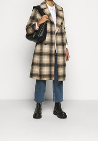 Cappotto a plaid in tonalità beige, nero e crema; silhouette oversized; abbinato a una camicia bianca e jeans blu; borsa nera; stivali neri chunky.