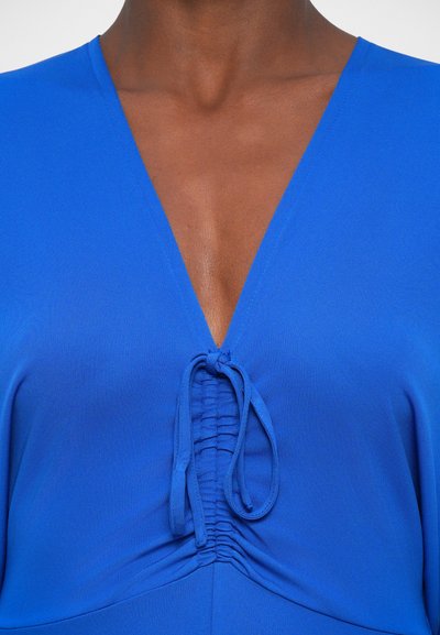 Modra tkanina s topom z globokim V-izrezom in nabranimi podrobnostmi na sredini. Gladka tekstura in nastavljiva vrvica pri izrezu.