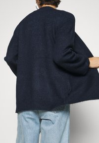 Granatowy sweter dziany o luźnym kroju, z zaokrąglonym dekoltem i rękawami balonowymi. Miękka tekstura z delikatnym połyskiem, noszony na jasnoniebieskich dżinsach.