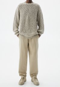 Personne portant un pull en maille grosse beige, un pantalon droit beige, des bottines à lacets beige, et un collier fin en chaîne argentée, les mains dans les poches.