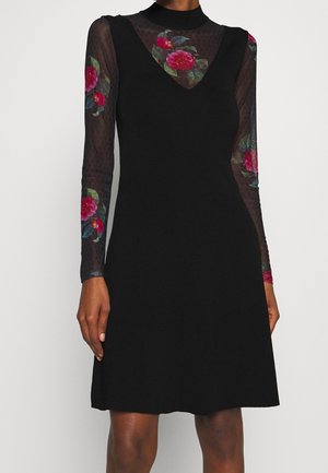 Woman wearing a black sleeveless dress over a sheer long-sleeve top with red and pink floral patterns on sleeves and chest.