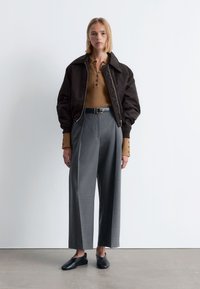 Braune Bomberjacke, beige gerippter Pullover mit Knopfleiste, graue weit geschnittene Hose und schwarze Slipper. Mattes Material und taillierte Passform.