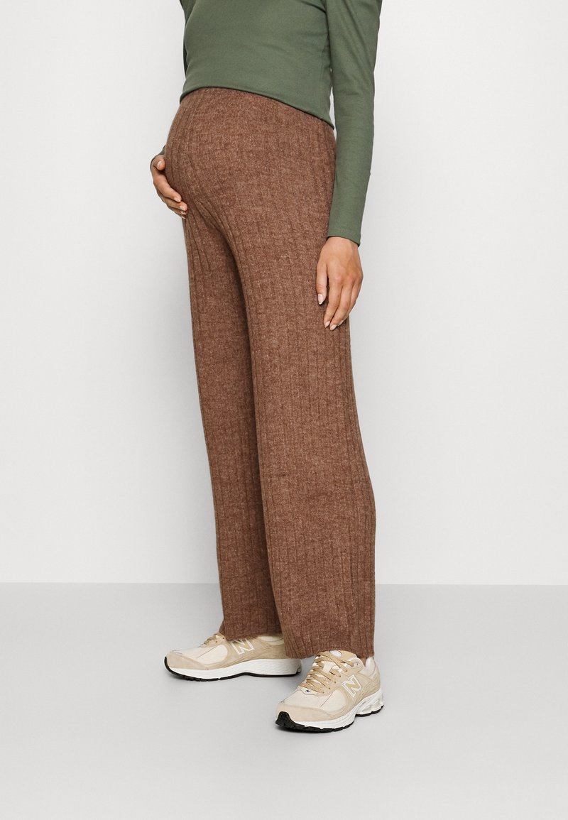 Personne enceinte portant un pantalon large en maille côtelée marron, un haut à manches longues vert olive et des baskets beiges, debout de profil avec une main sur le ventre.