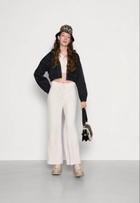 Jaqueta de denim preta oversized, top curto branco, calças largas cor creme, sapatilhas bege e um chapéu de balde com padrão de leopardo, segurando uma bolsa.