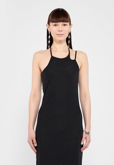 Femme aux cheveux bruns raides avec une frange, portant une robe noire sans manches avec de fines bretelles et de longues boucles d'argent sur un fond uni.
