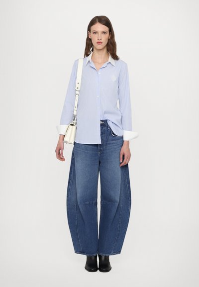 Light blue striped button-down shirt with white cuffs, paired with wide-leg blue jeans. Model holds a small silver handbag and wears black boots.