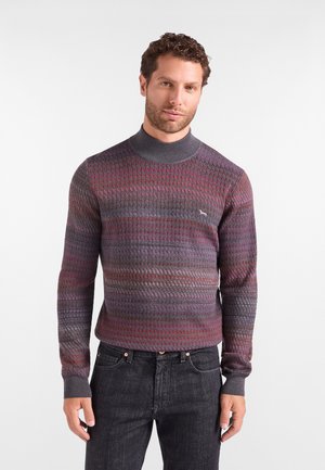 Uomo che indossa un maglione aderente in maglia strutturata con collo alto nelle tonalità di grigio, viola e rosso, abbinato a jeans neri.