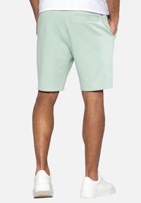 Shorts vert clair en mélange de coton avec poches latérales, ceinture élastique et ourlet cousu. Portés avec des baskets blanches.