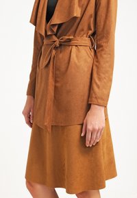 Femme portant une veste cache-cœur en daim marron avec une ceinture nouée et une jupe en daim marron assortie sur un fond blanc uni.
