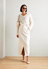 Pull en maille blanc avec un col en V, associé à une jupe en jean blanche arrivant aux genoux et comportant une fente à l'avant. Chaussures de couleur claire et lunettes de soleil.