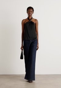 Black halter top with a knot detail, worn with wide-leg dark denim jeans. The model carries a small black handbag, standing against a neutral backdrop.