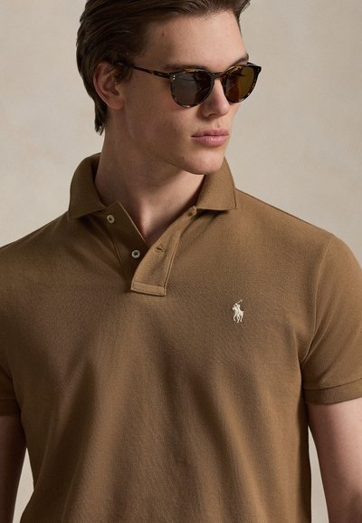 Καφέ πόλο πουκάμισο με υφή, ανοίγμα δύο κουμπιών και μικρό λευκό κεντητό λογότυπο. Το μοντέλο φορά γυαλιά ηλίου χελώνας.