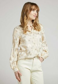 Blouse crème avec un motif floral, présente des accents métalliques. Manches longues avec poignets à boutons et un design à boutons sur le devant.