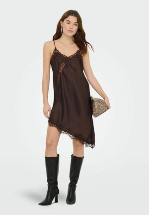 Žena u crnoj haljini s čipkastim rubom, crnim čizmama do koljena, držeći torbicu s uzorkom zmijske kože, stoji pred jednostavnom pozadinom.