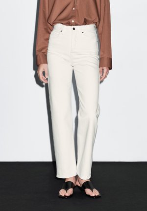 Femme portant un pantalon droit taille haute écru, une chemise marron à manches longues avec boutons, et des sandales noires ouvertes à l'avant.