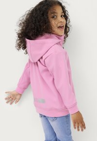 Rosa Kapuzenpullover mit weicher Textur, mit einem grauen Logodetail auf der linken Seite. Getragen mit hellblauen Jeans.