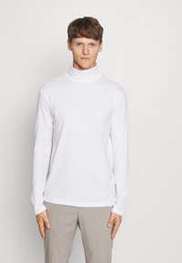 Suéter blanco de manga larga y cuello alto, hecho de una tela suave, con un diseño ajustado y cuello sin costuras. Combinado con pantalones de color claro.