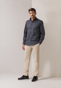 Marineblauw overhemd met knopen en een borstzak, gecombineerd met beige broek en zwarte sneakers. Het overhemd heeft een gestructureerd patroon en lange mouwen.