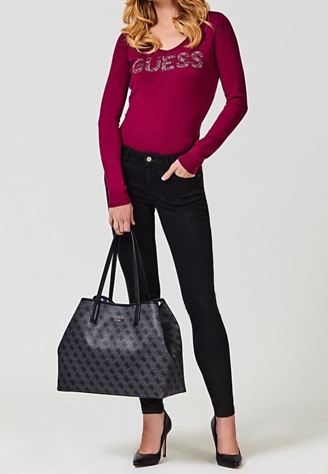 Femme portant un haut bordeaux à manches longues avec le texte ""GUESS"", jean skinny noir, talons noirs, tenant un grand sac cabas gris à motifs par les anses.
