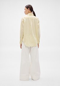 Dzeltena garroku krekls ar svītrām un apkakli, ar pogātām aprocēm, kopā ar plašām baltām biksēm, kas izceļ brīvu piegriezumu un auduma tekstūru.