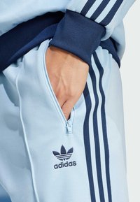 Training suit Adidas de culoare albastru deschis, cu trei dungi albastru închis și buzunar cu fermoar. Prezintă manșete ribbate și logo brodat pe piept.
