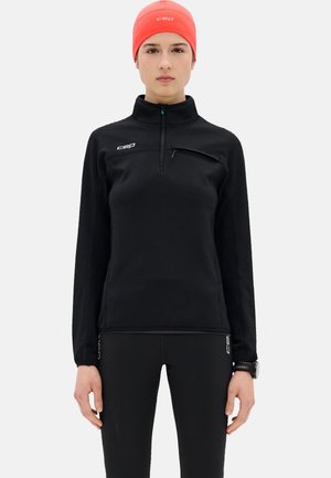 Vrouw draagt een zwarte sporttop met lange mouwen met een borstzak met rits, zwarte legging, een oranje muts en een zwart polshorloge.