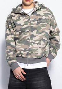 Beżowa, zielona i szara bluza z zamkiem błyskawicznym w kamuflażu. Posiada logo z przodu, kaptur i boczne kieszenie. Rybakowane mankiety i dół. Gładka struktura materiału.