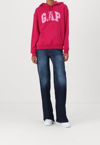 Osoba v jasně růžovém mikině GAP, tmavě modrých rovně střižených džínách a bílých teniskách, stojící na pozadí čistě bílé stěny.