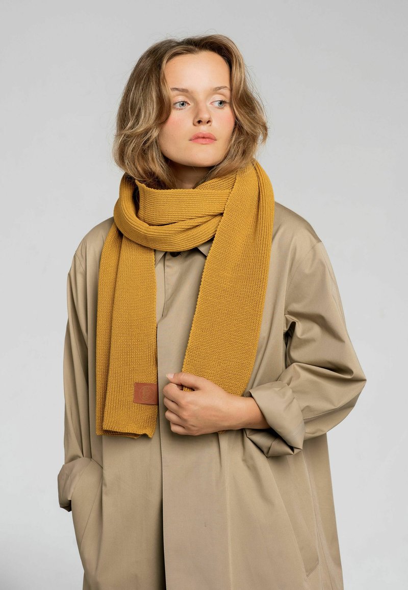 Musztardowożółty dziany szalik o teksturowanym wzorze, zarzucony na szyję, w połączeniu z jasnobeżowym płaszczem z szerokimi rękawami.