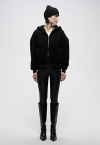 Crna teksturirana majica s kapuljačom i patentnim zatvaračem, nošena preko bijele majice, uparena s crnim tajicama i šiljastim crnim čizmama. Crna pletena kapa na glavi.