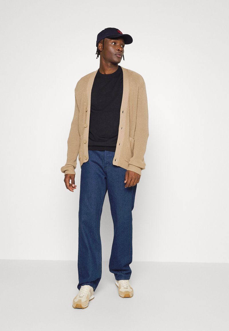 Man met een zwarte pet, een beige cardigan over een zwart shirt, blauwe jeans en beige sneakers die tegen een witte achtergrond staat.